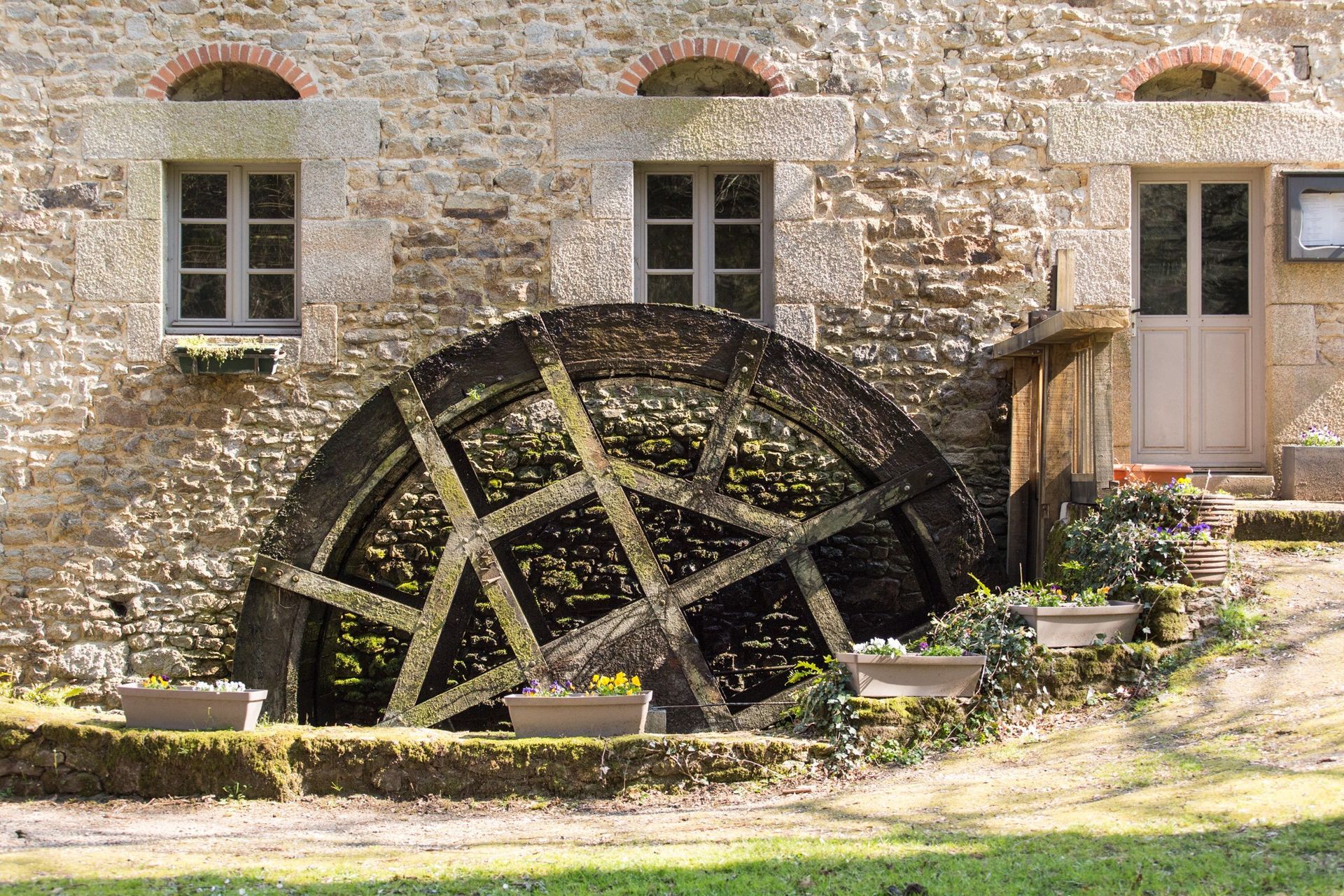 Une roue à aubes usée par le temps se dresse devant un bâtiment en pierre avec des fenêtres et une porte ; journée ensoleillée.