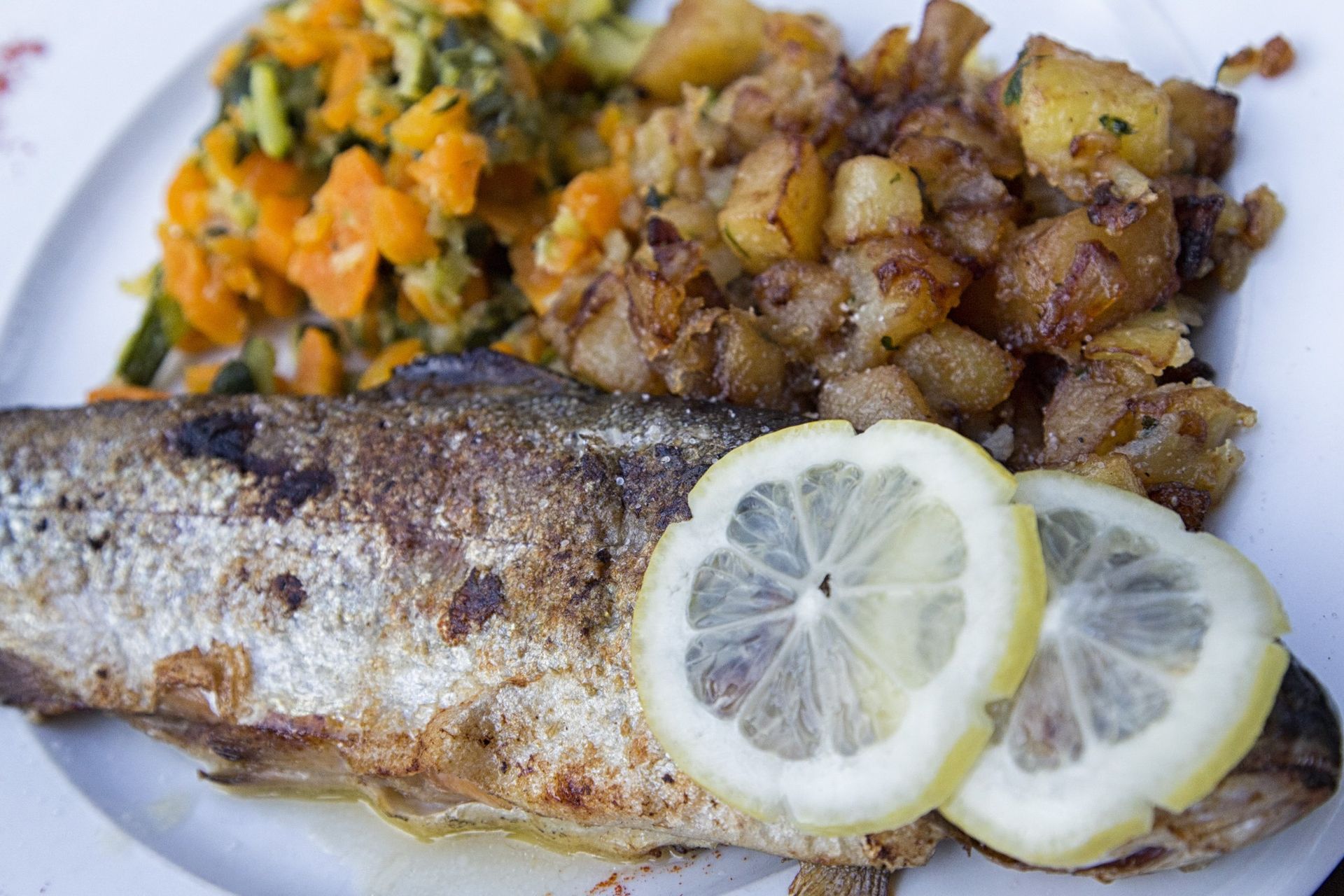 Poisson poêlé avec des tranches de citron, des pommes de terre et un mélange de légumes sur une assiette blanche.