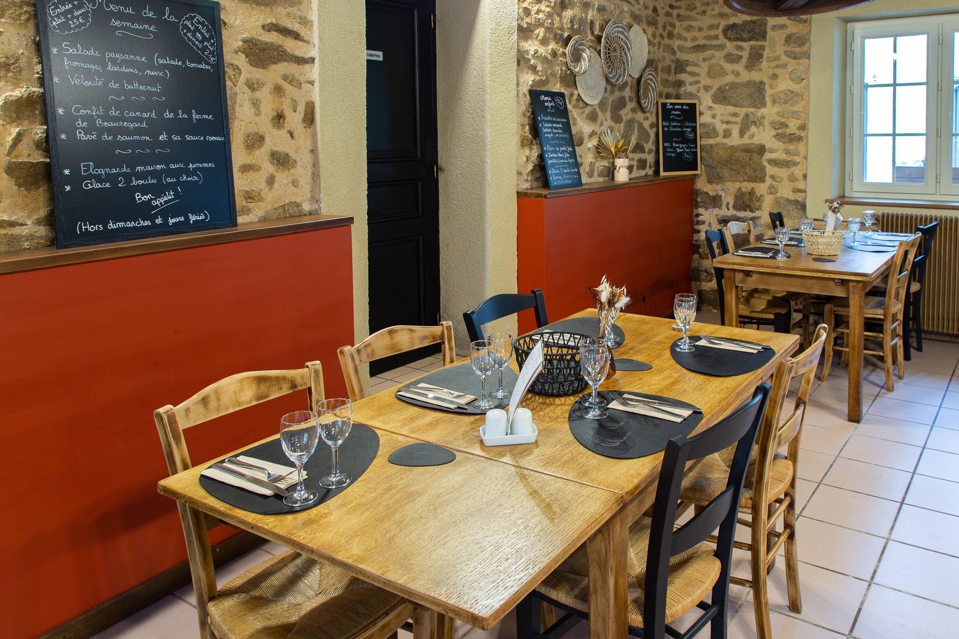 Intérieur du restaurant avec des tables en bois dressées pour les repas, des murs en pierre et des ardoises avec les menus.