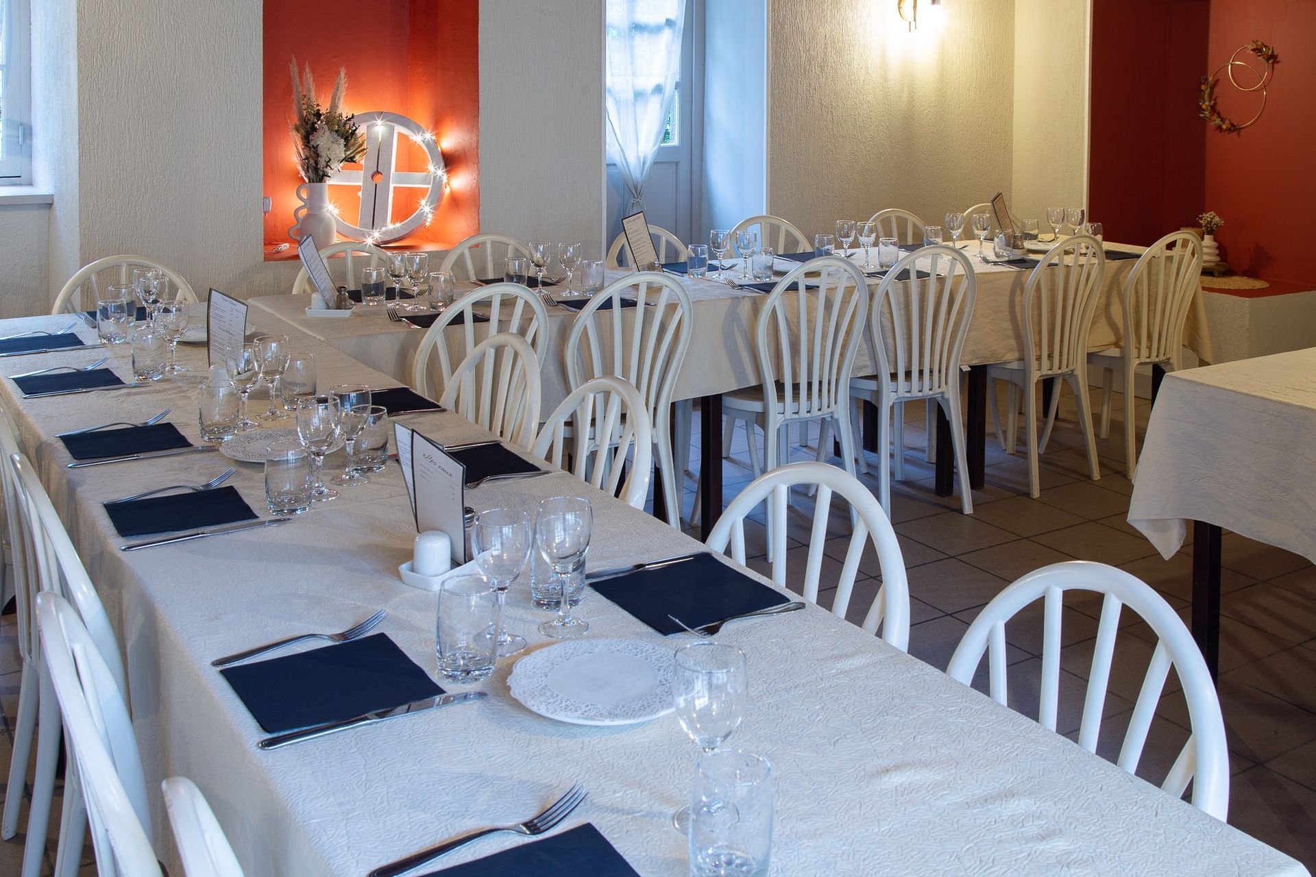 Salle à manger dressée pour un repas. Tables nappées de blanc, chaises blanches et couverts. Murs rouges et blancs.