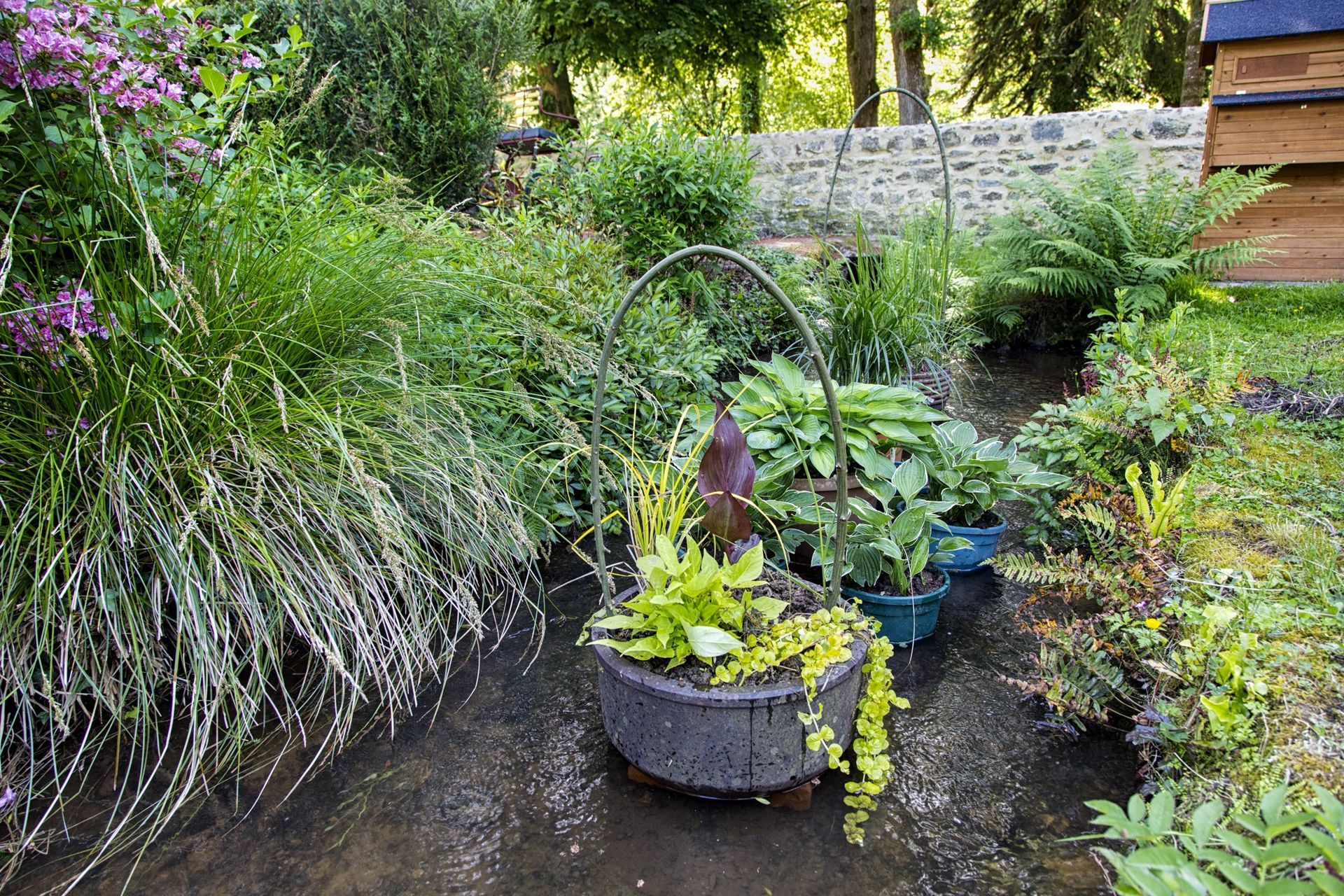 Un panier en pierre rempli de verdure est installé dans un ruisseau, entouré de plantes luxuriantes et d'un muret en pierre.