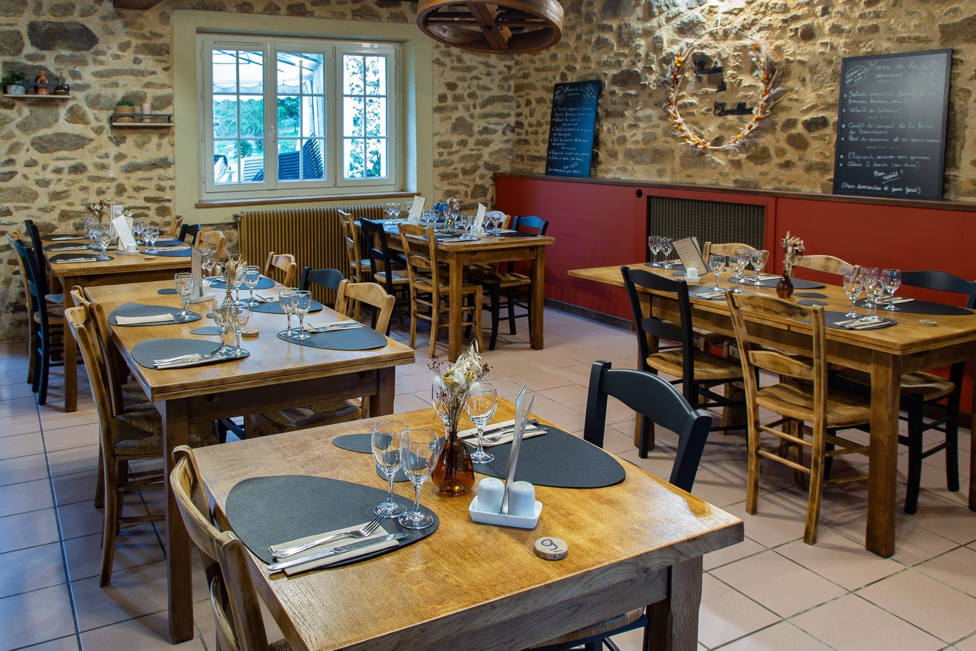 Intérieur d'un restaurant avec tables et chaises en bois, murs en pierre et une fenêtre. Les tables sont dressées avec des couverts et des verres.