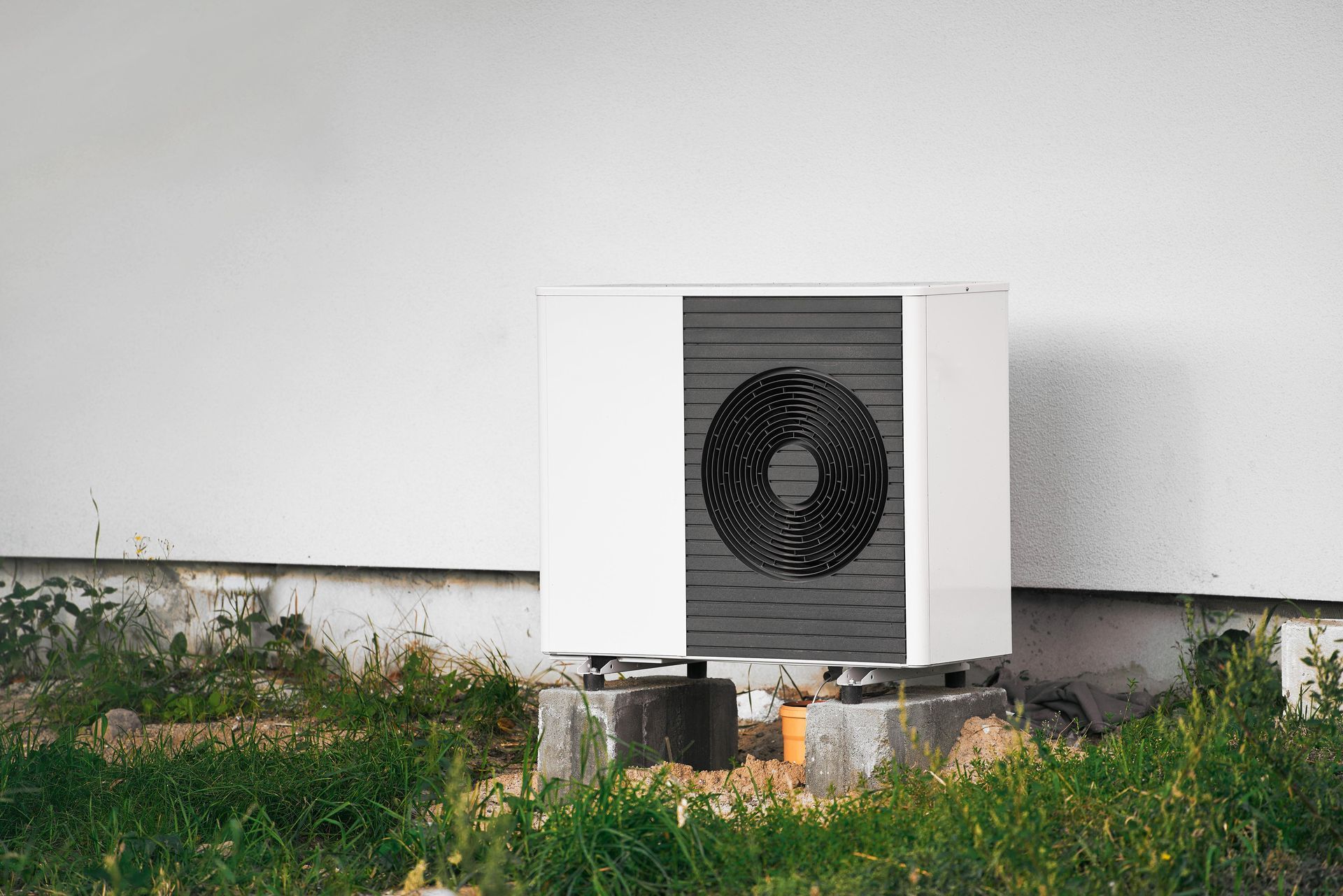 Weiße rechteckige Klimaanlage vor einer weißen Wand, auf einem Betonsockel im überwucherten Gras.