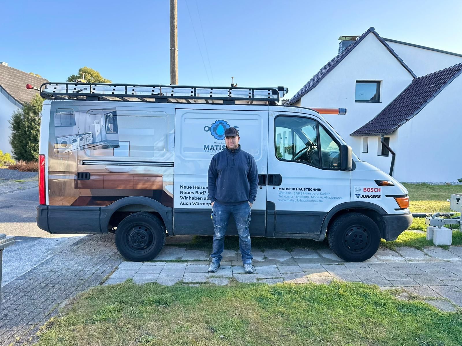 An einem sonnigen Tag steht ein Mann neben einem weißen Lieferwagen mit Firmenlogo und holzähnlichem Design vor einem Haus.
