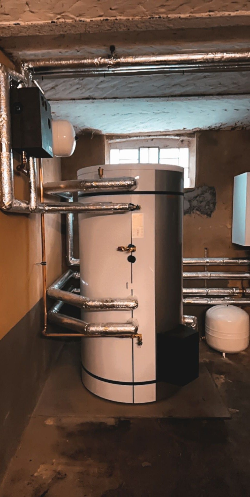 Ein Warmwasserbereiter in einem Keller mit isolierten Rohren, einem weißen Ausdehnungsgefäß und einem Fenster.