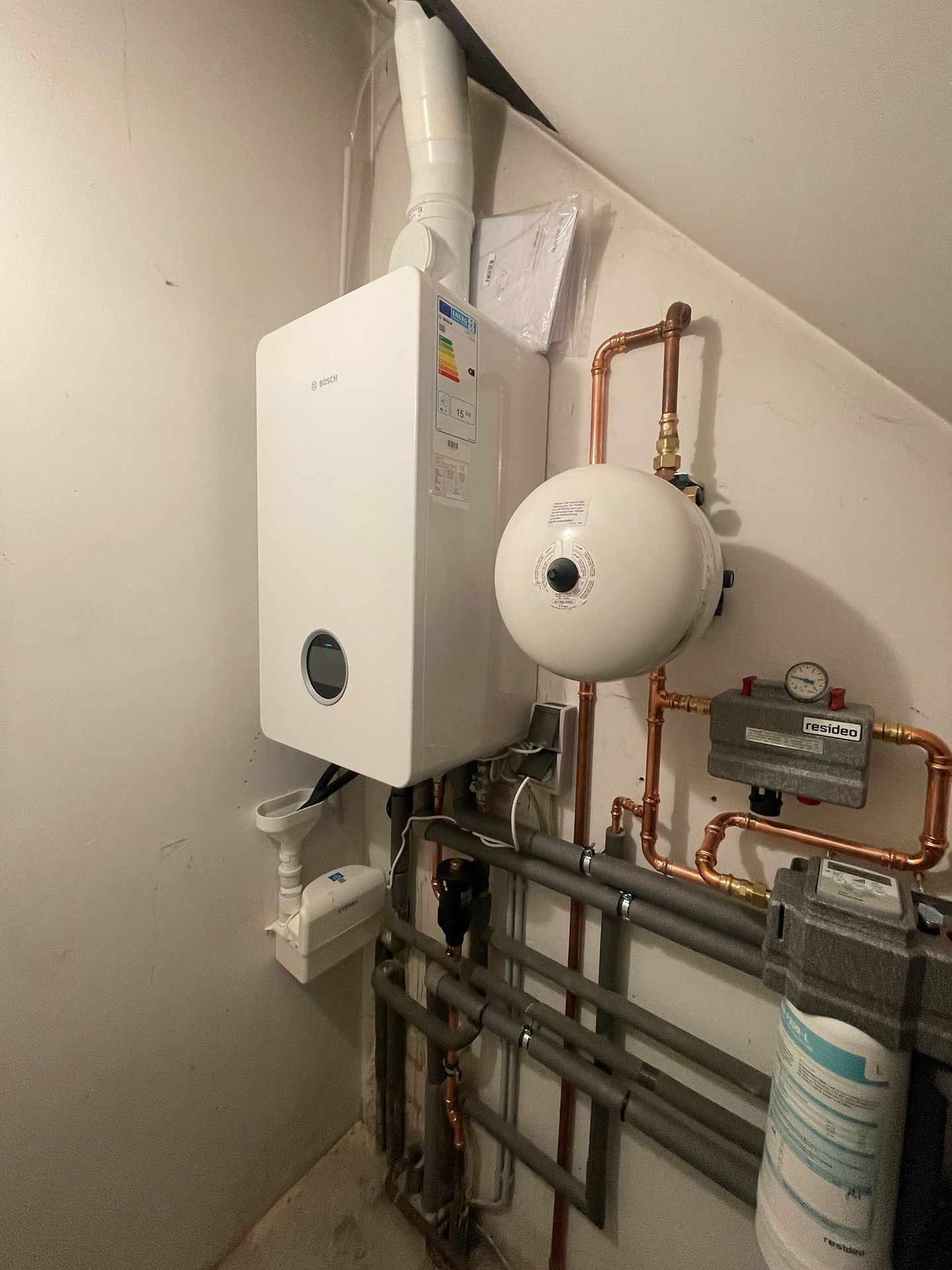 Weißer Heizkessel und angeschlossene Geräte in einem Hauswirtschaftsraum, mit Kupferrohren und einem weißen Tank.