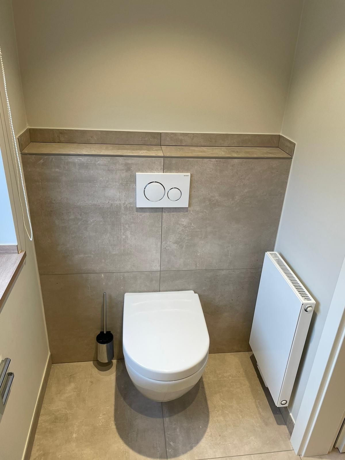 Eine moderne Toilette in einem kleinen, beige gefliesten Raum, mit Bürste und Heizkörper.