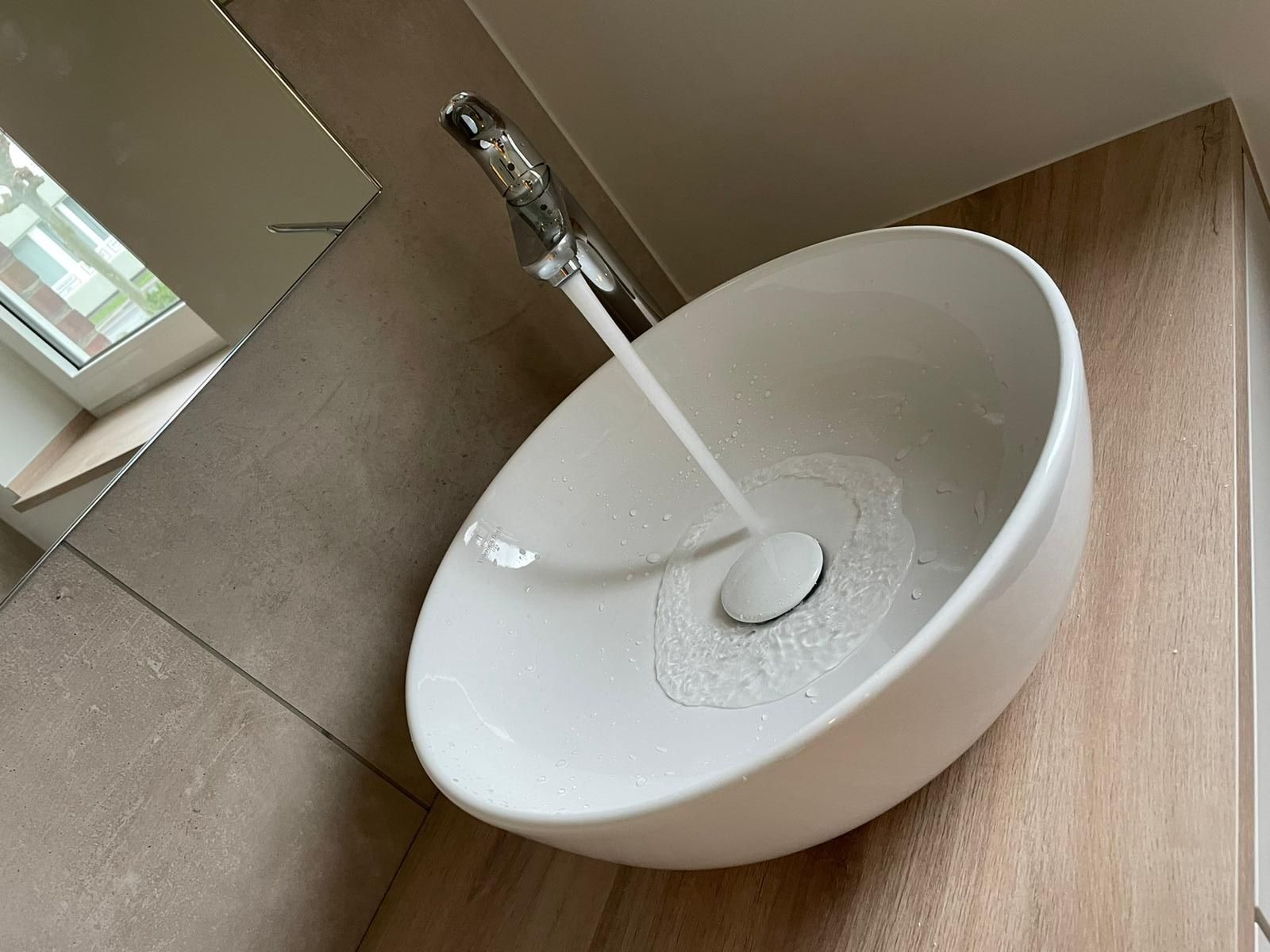 Weißes Waschbecken mit fließendem Wasser aus einem Chromhahn, vor einer Arbeitsplatte aus hellem Holz.