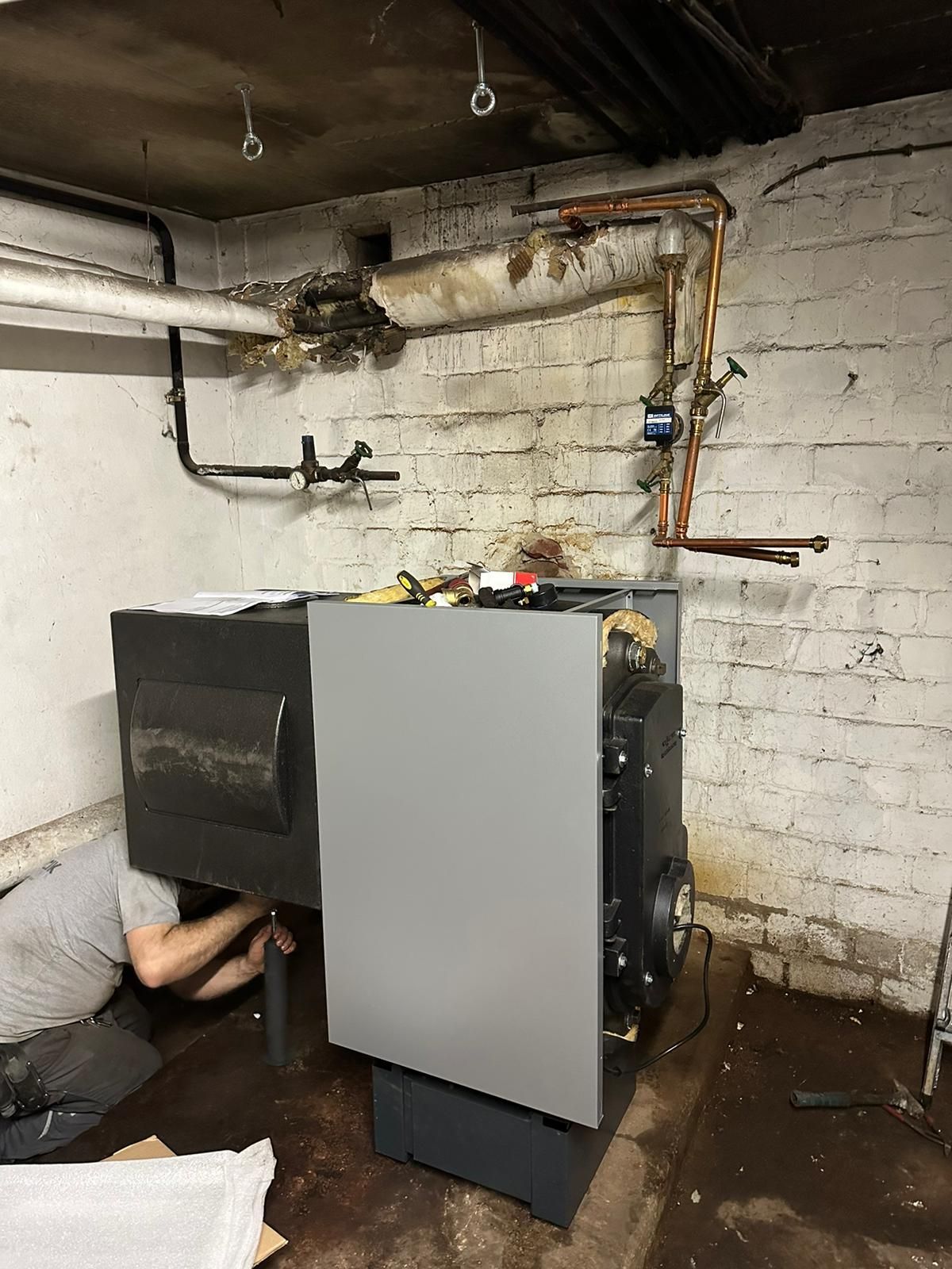 Person, die an einem Heizkessel in einem Keller mit freiliegenden Rohren und einer Ziegelwand arbeitet.