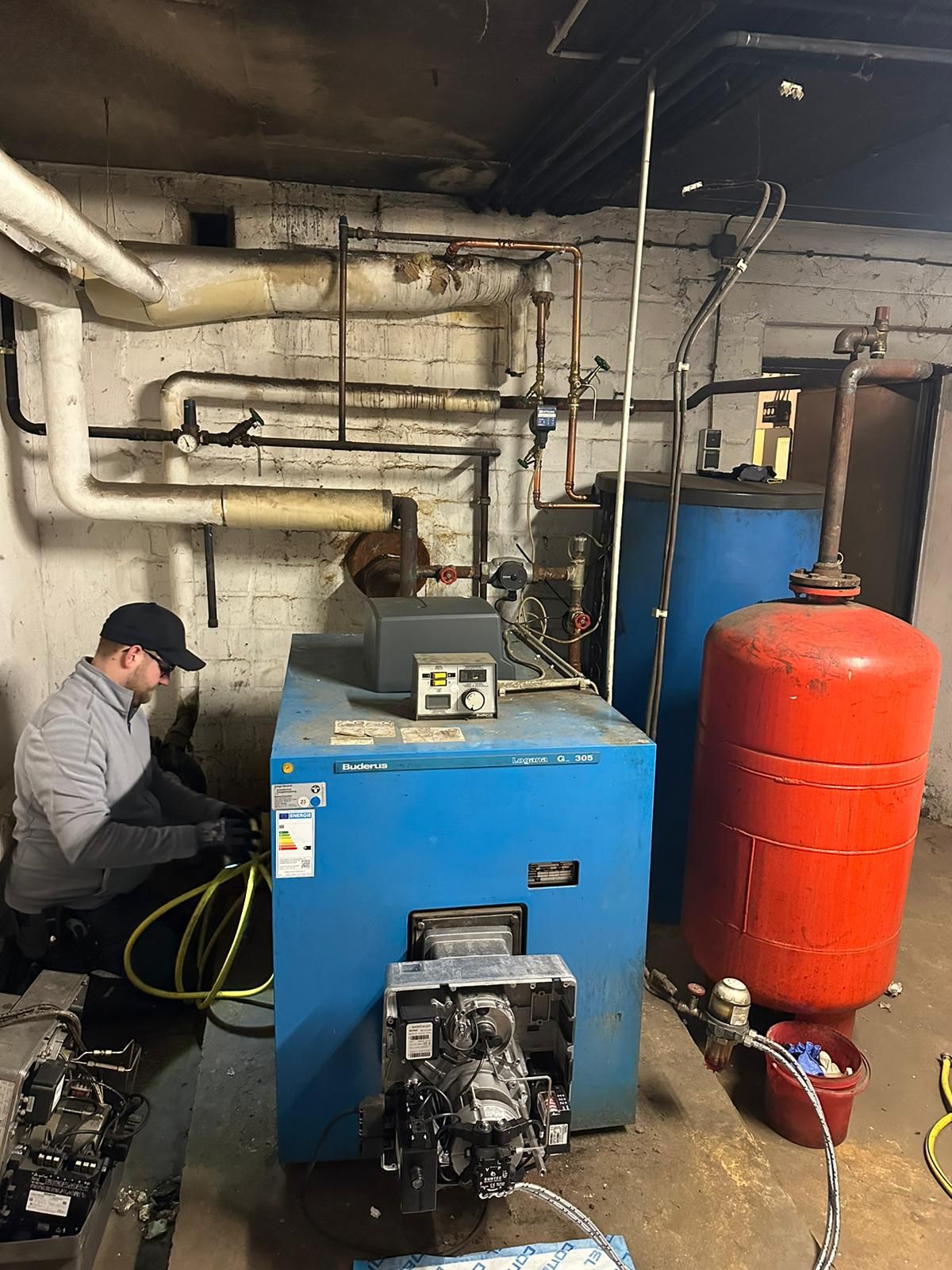 Mann arbeitet an einem blauen Heizkessel im Keller. Rohre, Tanks und Werkzeuge sind sichtbar.