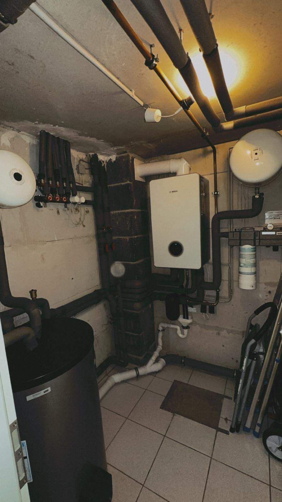 Keller mit Heizungsanlage: Wassertank, Boiler, Rohre und Fliesenboden.