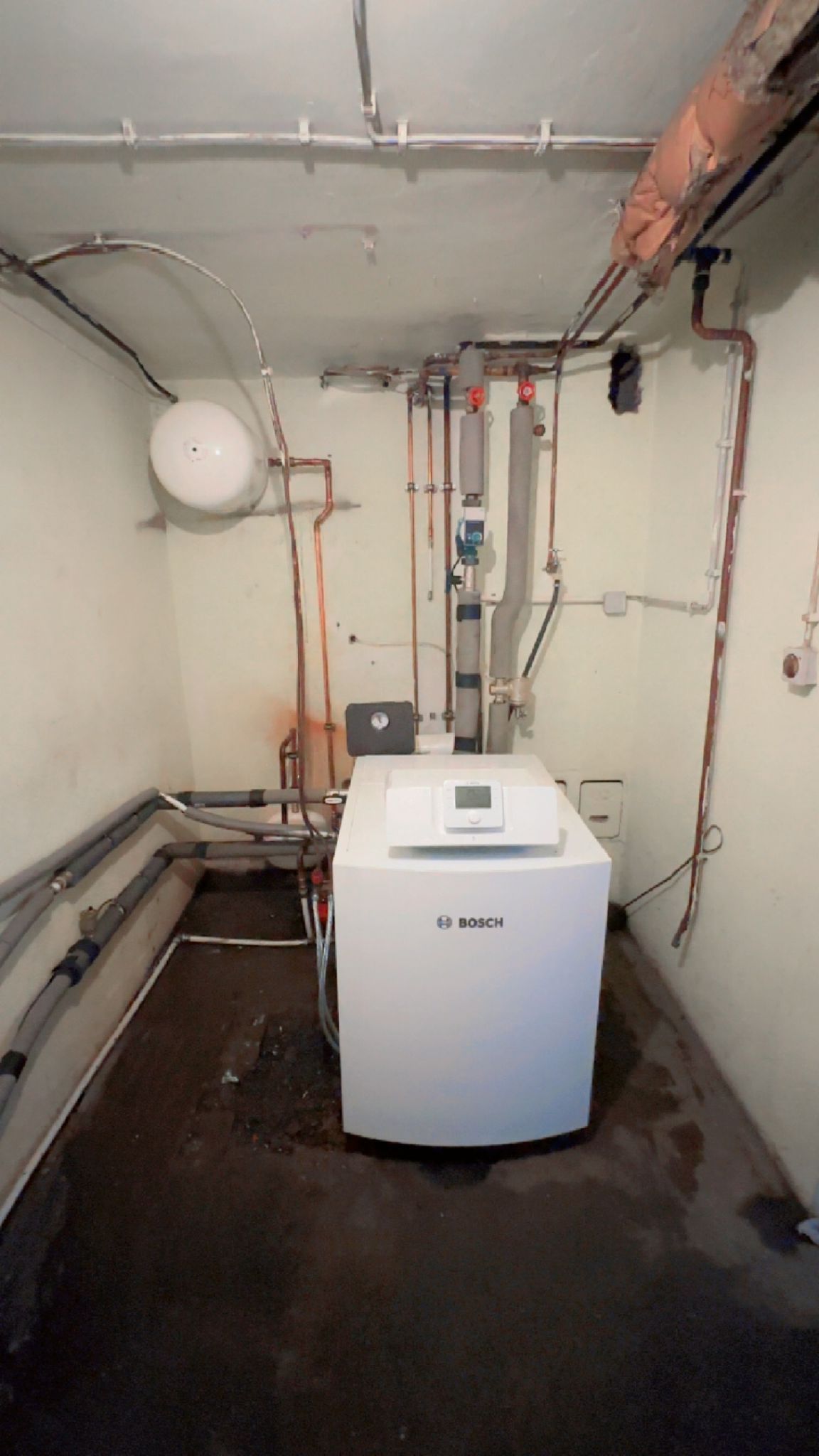 Ein Hauswirtschaftsraum mit einem weißen Heizkessel, Rohren und einem weißen Ausdehnungsgefäß an der Wand.