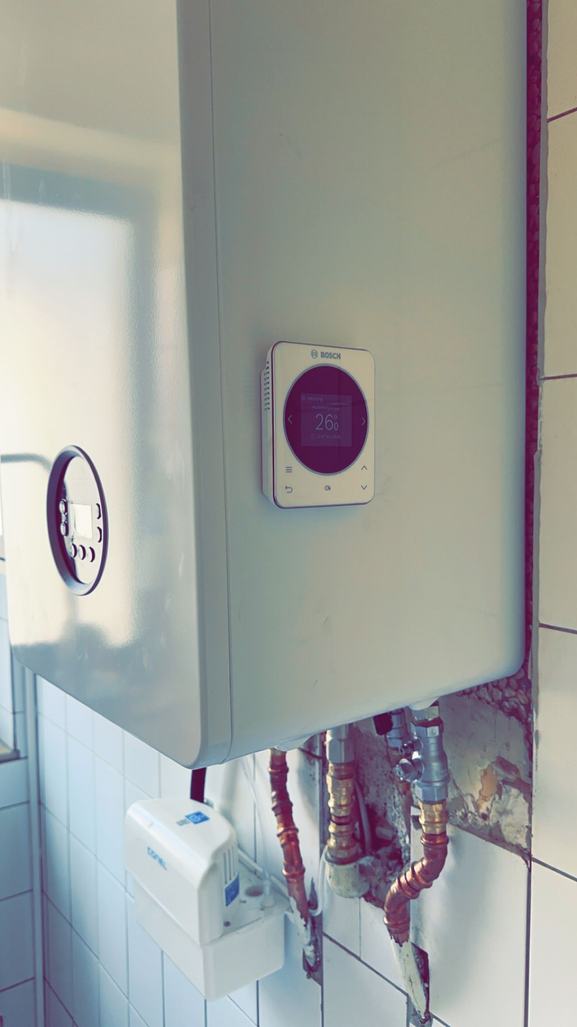 Weißer Warmwasserbereiter mit Kupferrohren, an einer gefliesten Wand montiert. Ein kleiner Thermostat ist sichtbar.