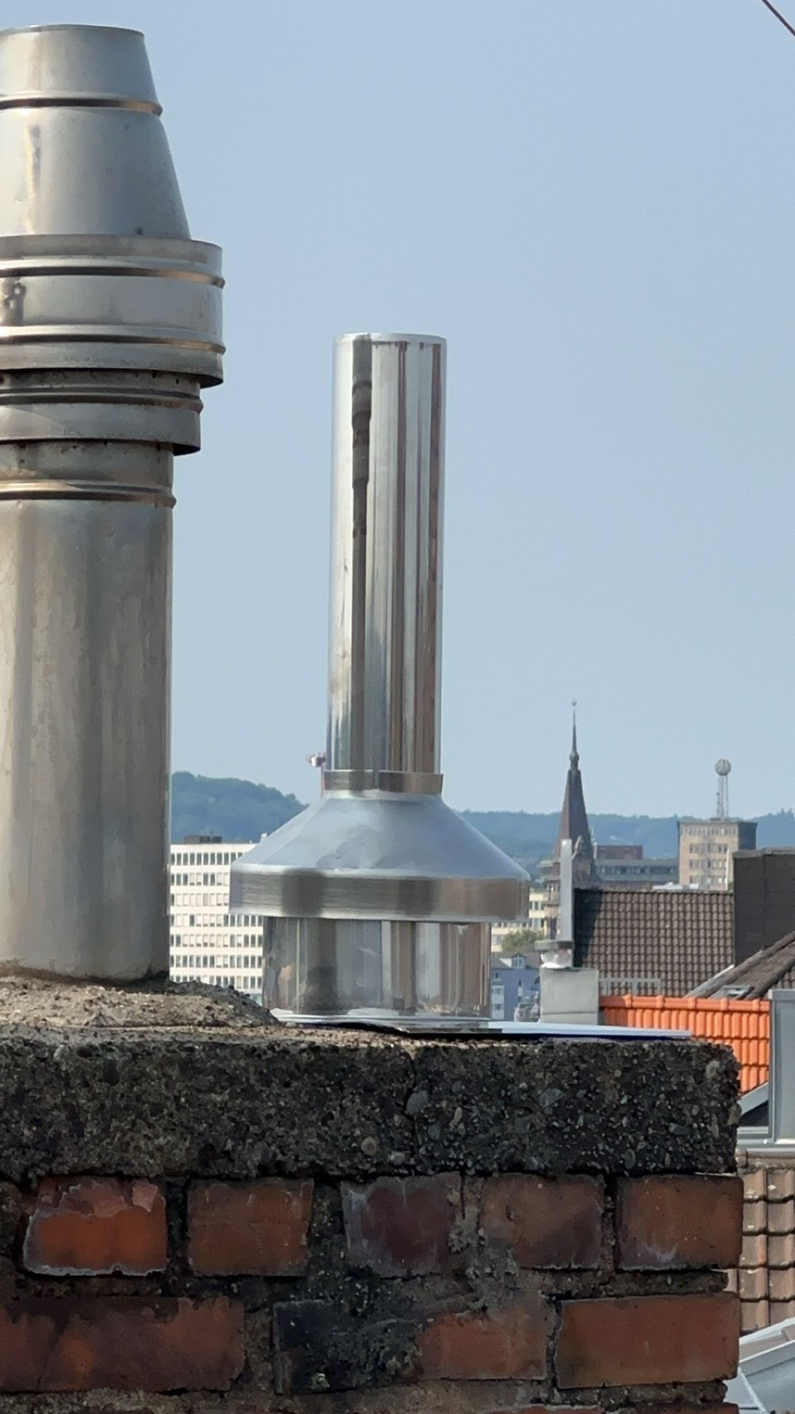 Glänzendes Kaminrohr aus Metall auf einem gemauerten Schornstein, Hintergrund die Skyline der Stadt.