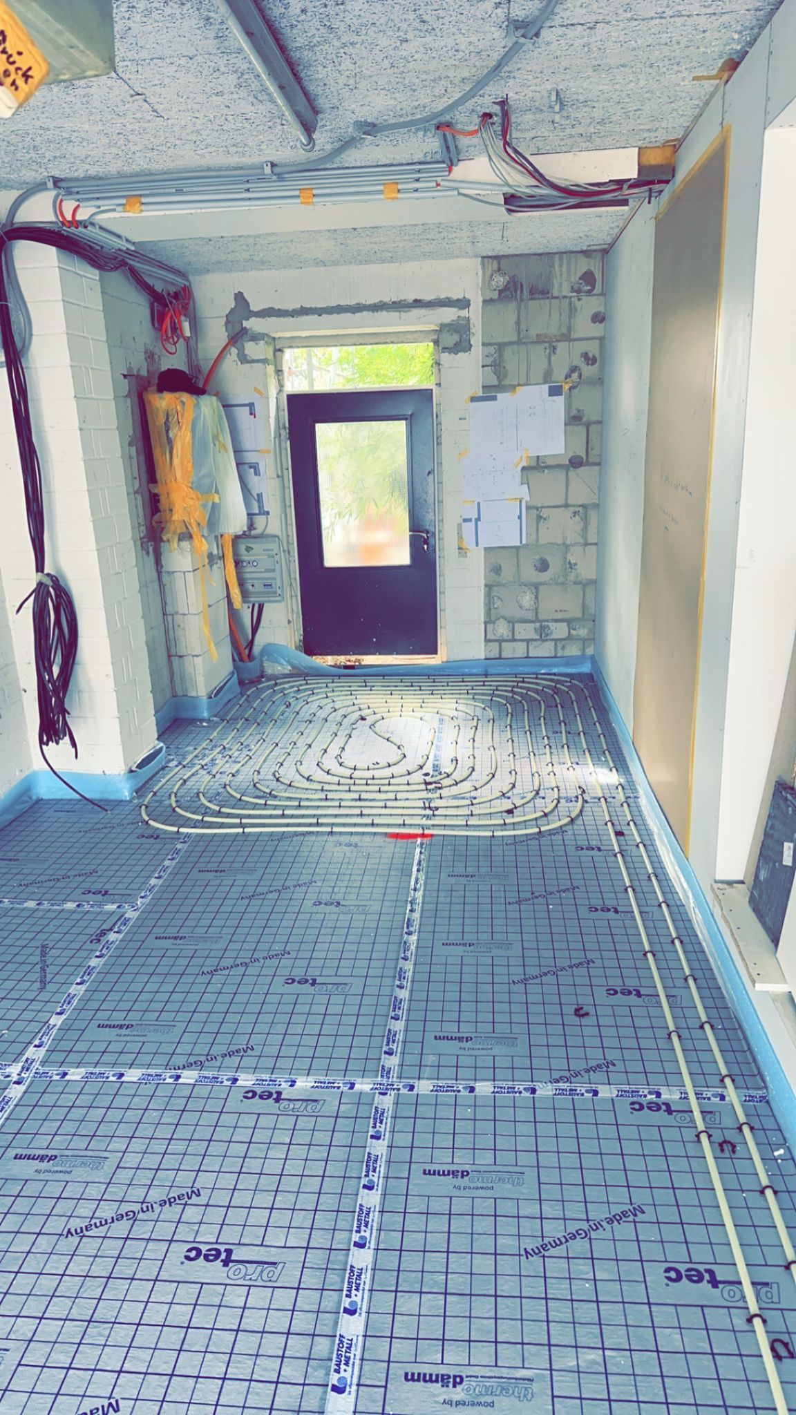 Flur auf einer Baustelle mit blauem Bodenbelag, offener Tür, Verkabelung an den Wänden und gelben Baugeräten.