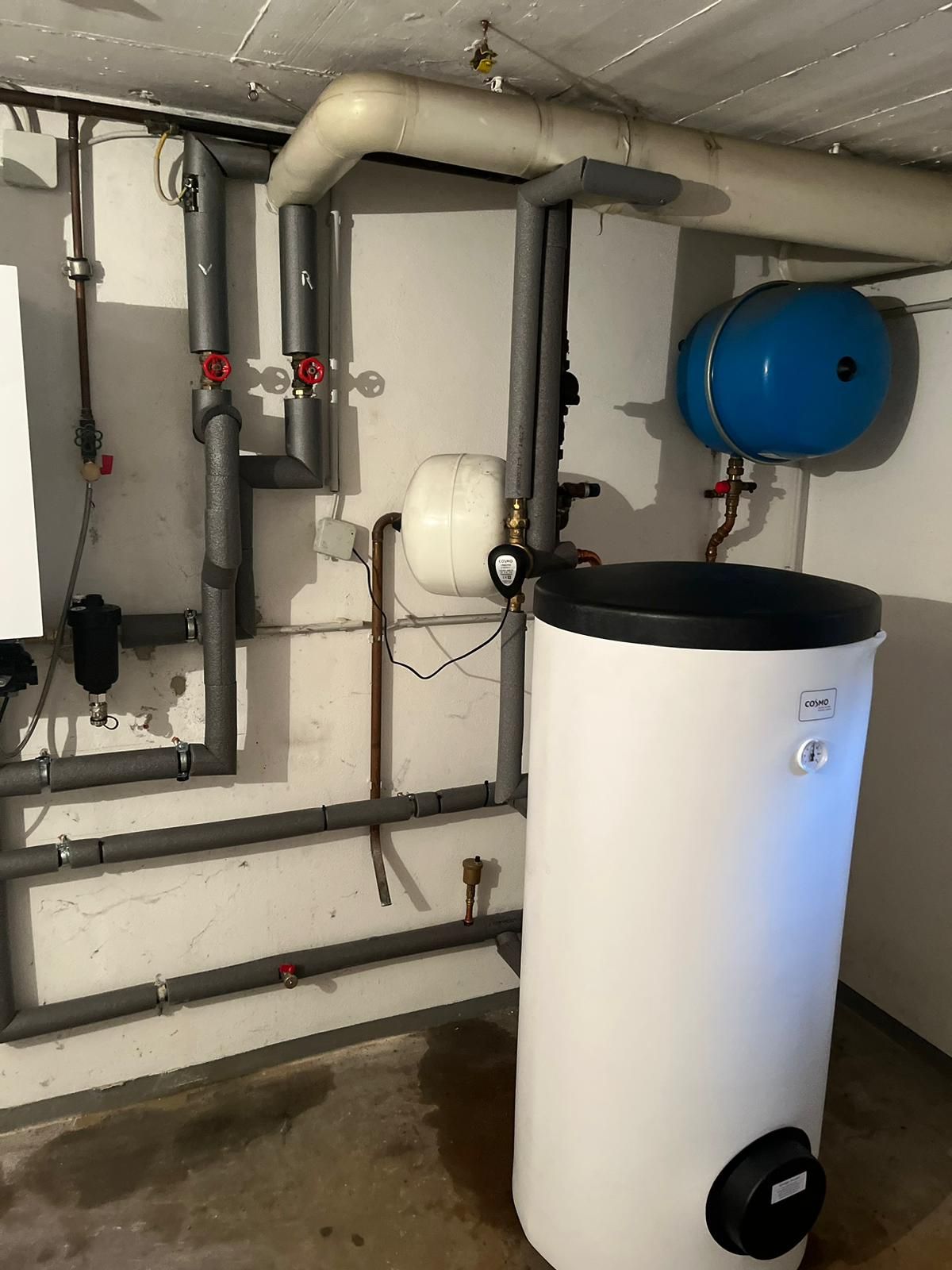 Sanitärsystem mit isolierten Rohren, weißem Wassertank, blauem Ausdehnungsgefäß und verschiedenen Ventilen in einem Keller.