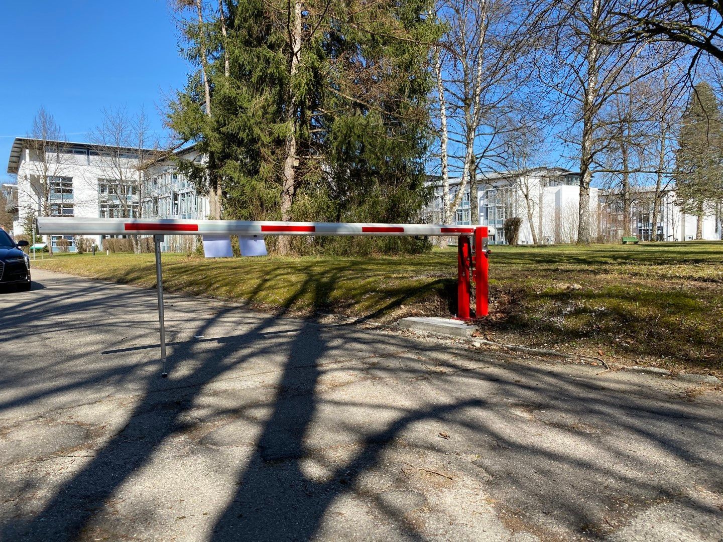 Ein rot-weißes Schrankentor sperrt eine Straße, im Hintergrund sind weiße Gebäude und Bäume zu sehen.