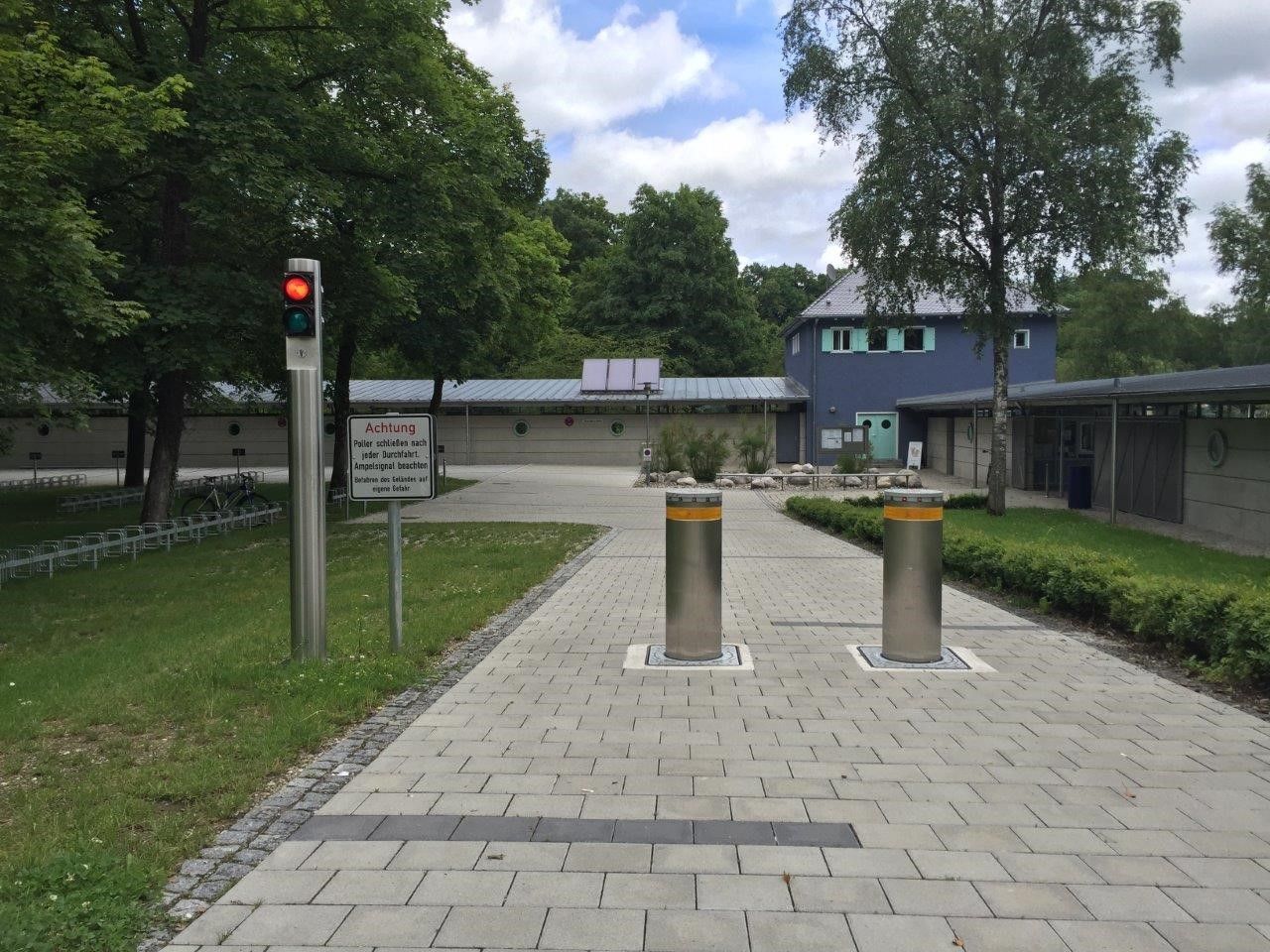 Eingang mit erhöhten silbernen Pollern und roter Ampel; im Hintergrund ein blaues Gebäude und Bäume.