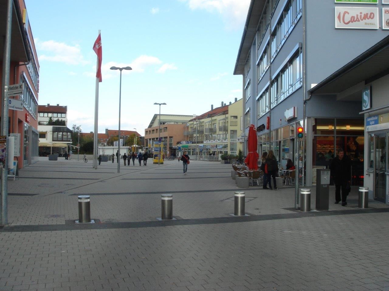 Ein Fußgängerplatz in einer europäischen Stadt mit Geschäften und Passanten. Graues Pflaster, Gebäude und Poller.