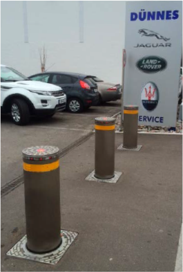 Drei braune Poller mit orangefarbenen Spitzen vor einem Autohaus, dahinter parken Autos.