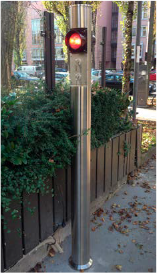 Rotes Fußgängersignal auf einem Edelstahlmast neben einem Holzzaun und Büschen.