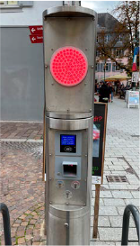Fahrradparkstation aus Edelstahl mit Rotlicht, Display und Bezahlschlitz. Im Außenbereich, im Hintergrund Fußgänger.