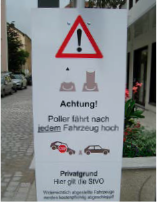 Schild: Warnung vor Pollern, Text, Symbole. Rotes Dreieck mit Ausrufezeichen, zwei Pollersymbole, Autosymbole.