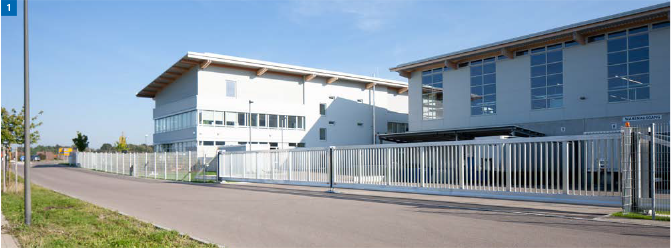 Zweistöckige helle Gebäude hinter einem weißen Zaun und einem Eingangstor an einem sonnigen Tag.