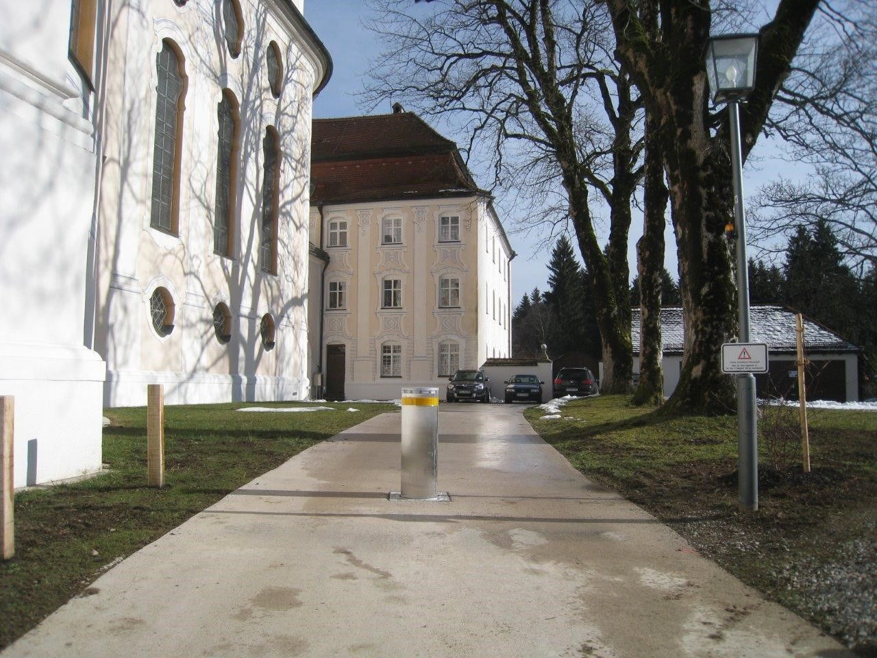Weg, der zu einem Gebäude führt, in dessen Ferne Autos geparkt sind, große Bäume und ein Lichtmast.