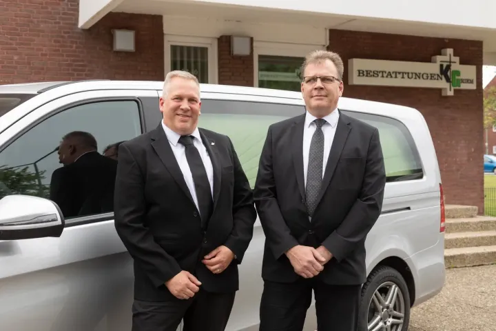 Zwei Männer in Anzug und Krawatte stehen vor einem Lieferwagen.