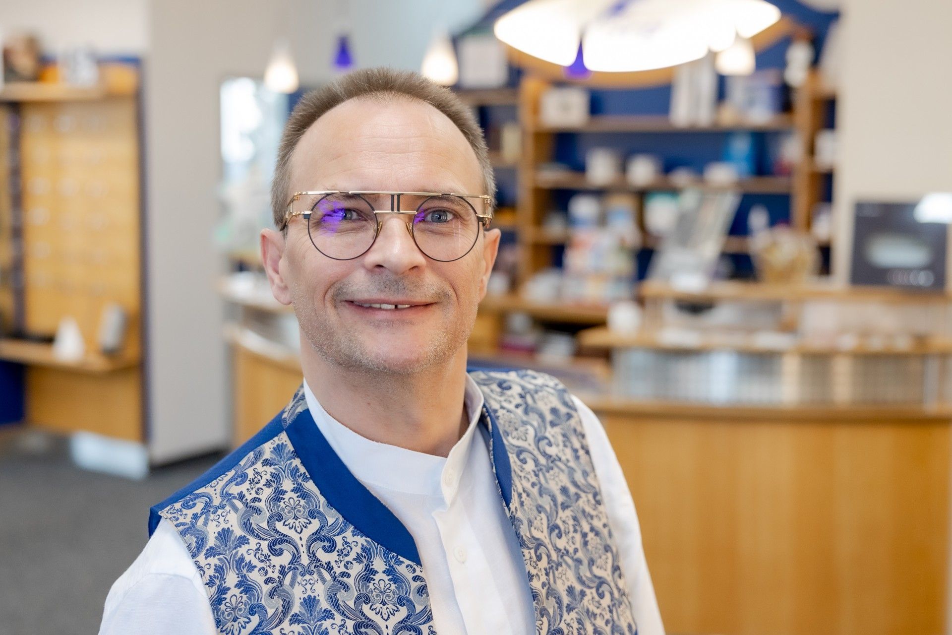 Ein Mann mit Brille und blauer Weste steht vor einem Tresen in einer Apotheke.