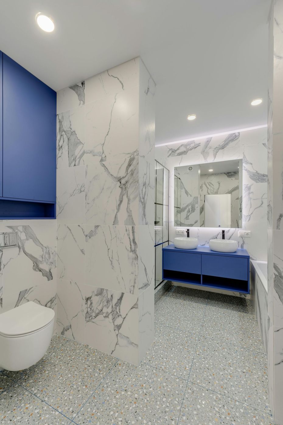 Baño moderno con paredes de mármol blanco, tocador azul, espejo y suelo de terrazo.