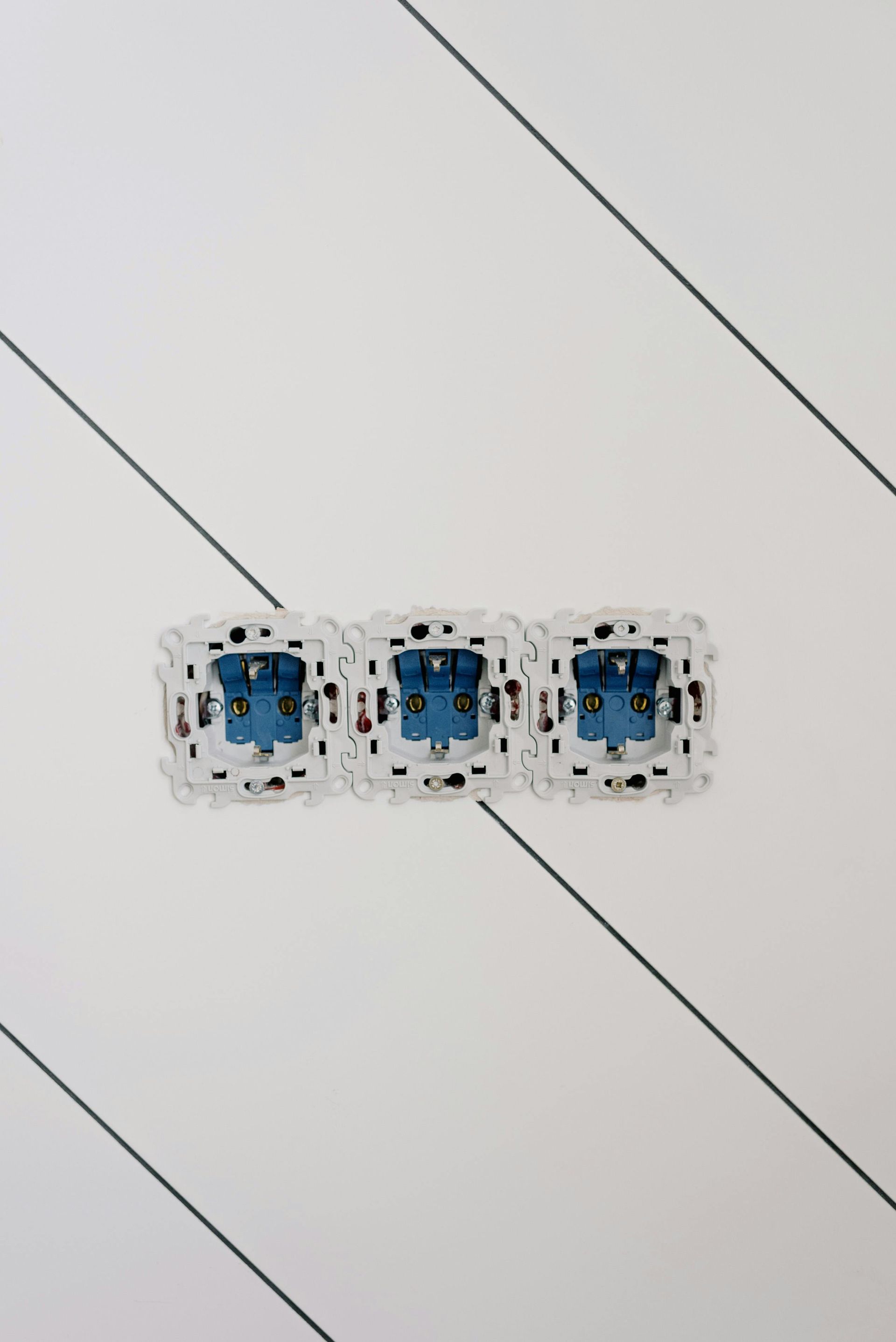 Tres enchufes eléctricos azules y blancos alineados en una pared clara con líneas oscuras diagonales.