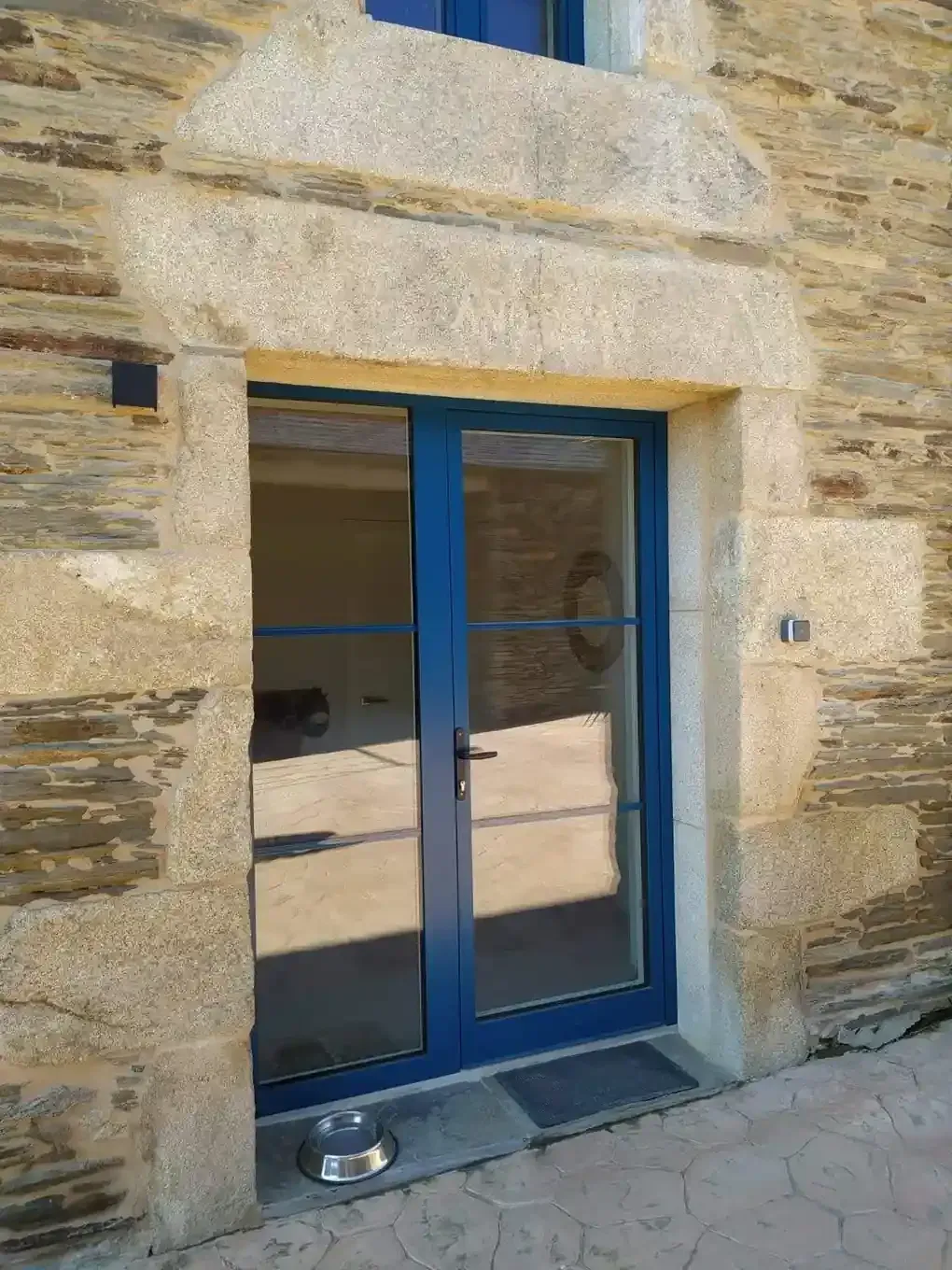 Puerta de cristal con marco azul en una pared de piedra, con un pequeño cuenco de metal en el umbral.