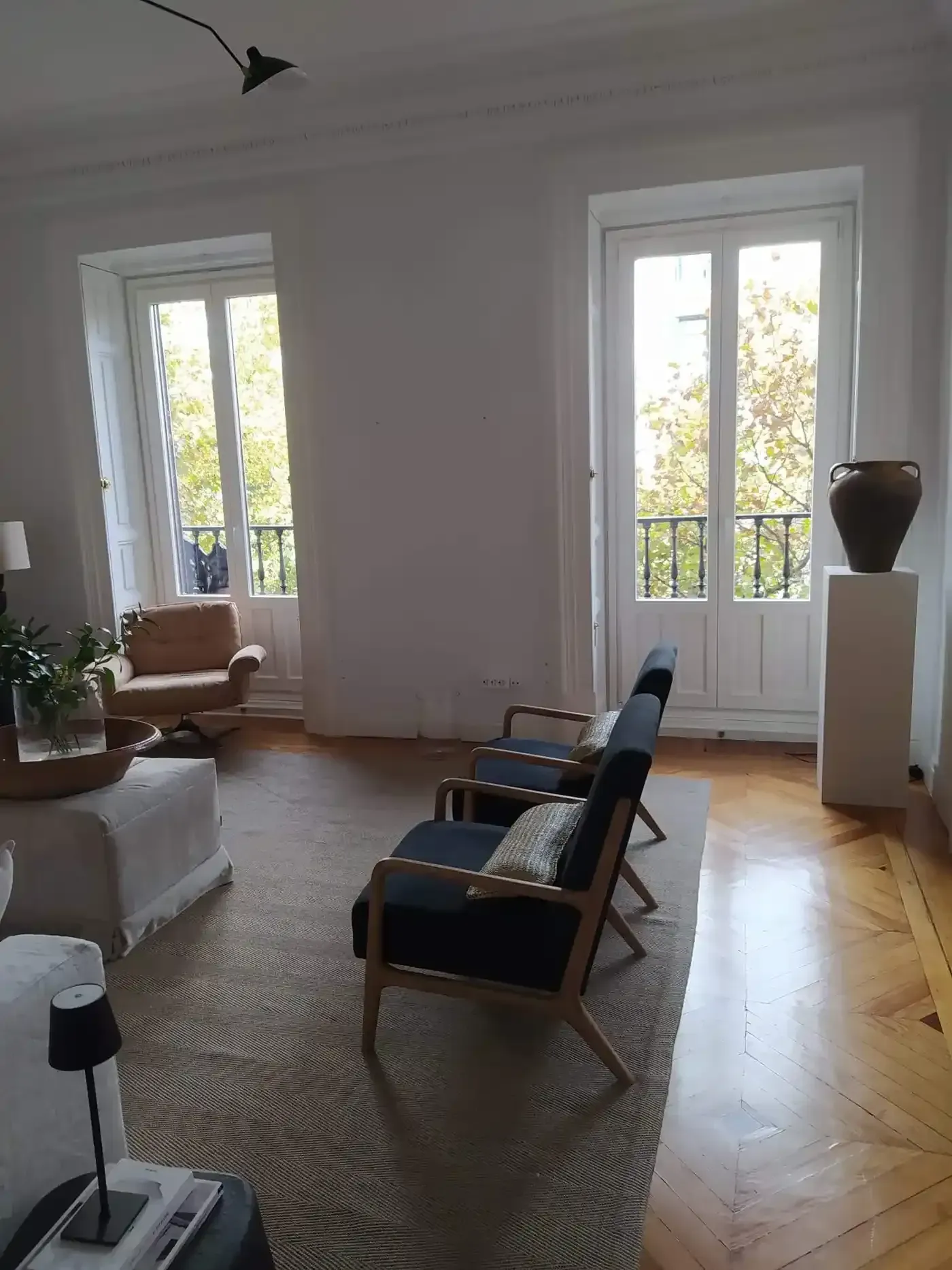 Sala de estar luminosa con grandes ventanales, suelo de madera en espiga y dos sillas azules sobre una alfombra tejida.