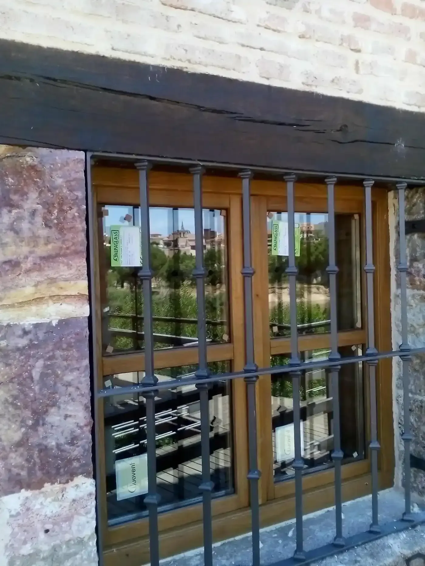 Ventana enrejada en un muro de piedra, que refleja los edificios y la vegetación del exterior.