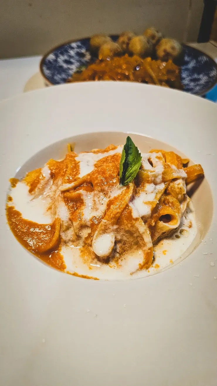 Plato blanco de pasta con salsa rojo anaranjada y albahaca, con otro plato borroso al fondo.