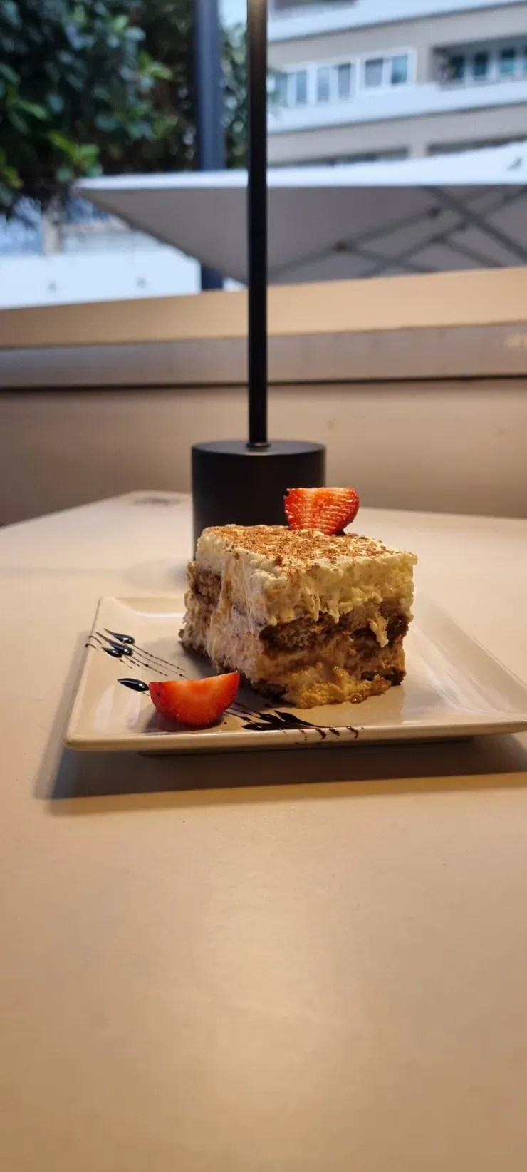 Pequeño pastelito de postre cubierto con fresas en un plato cuadrado junto a la ventana de una cafetería.