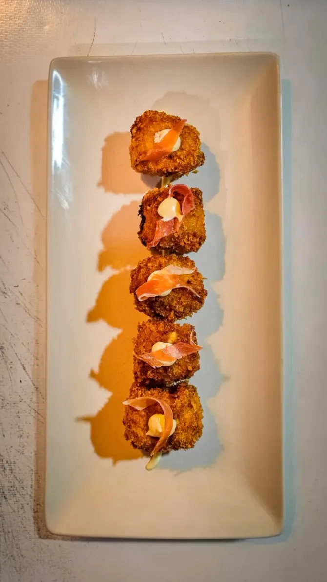 Cuatro aperitivos del tamaño de un bocado, dispuestos en línea vertical sobre un plato rectangular blanco alargado.