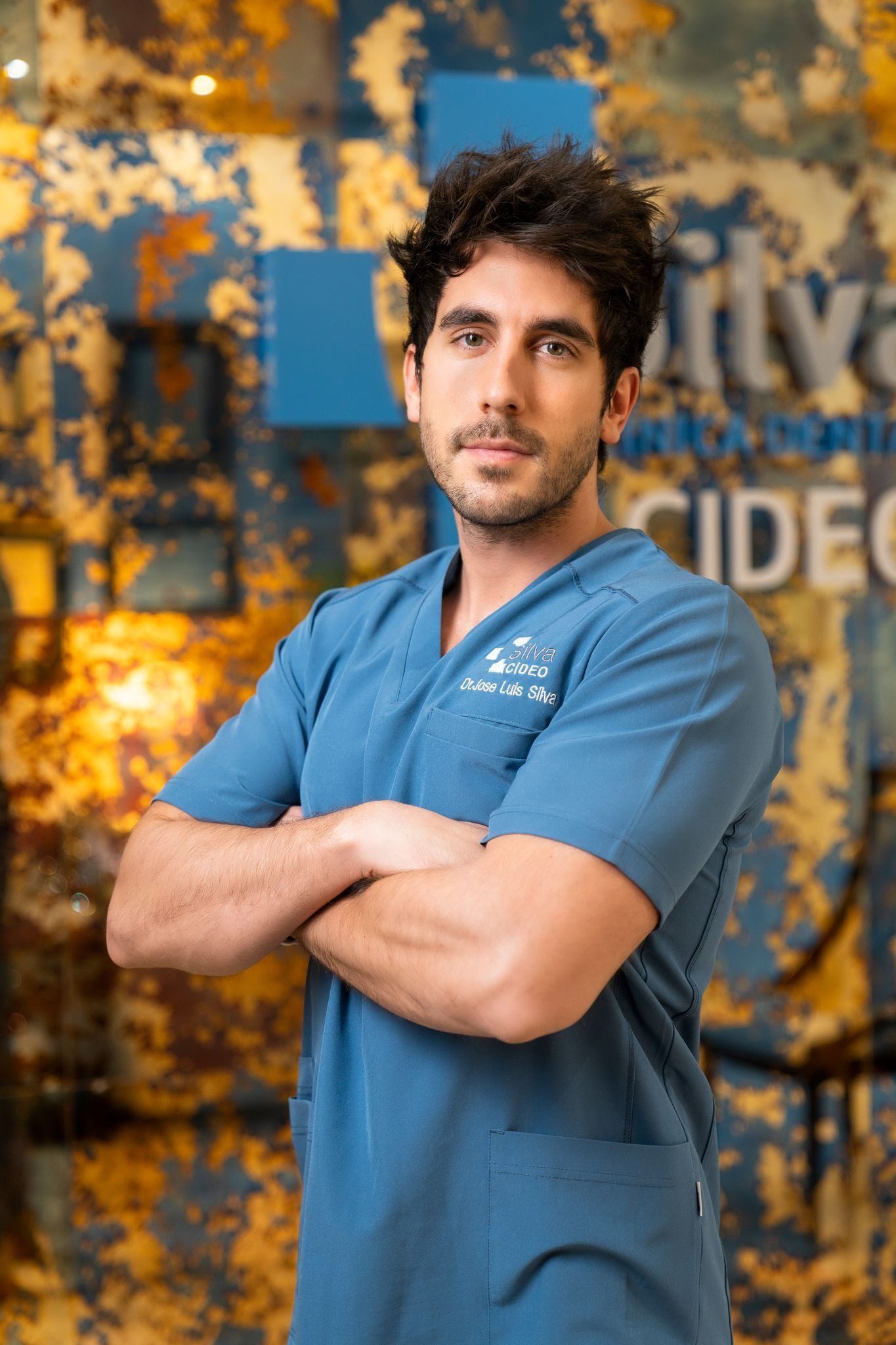 Hombre con uniforme azul y brazos cruzados, de pie en el interior.