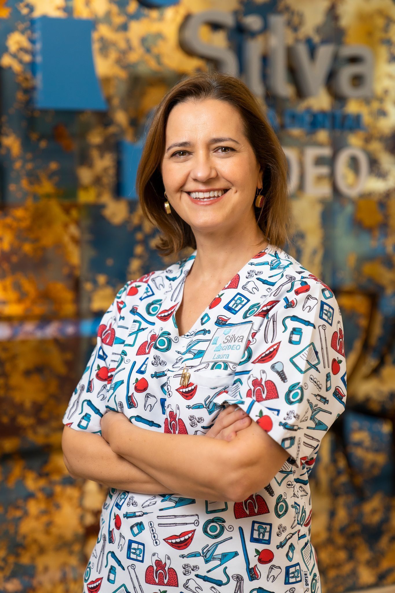 Una mujer con los brazos cruzados, vestida con un uniforme estampado, sonríe en un consultorio dental.