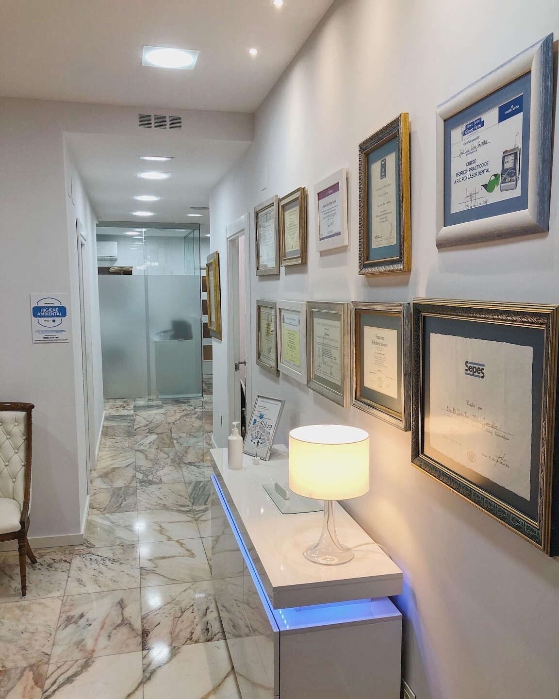 Pasillo largo en consultorio médico con piso de mármol, certificados enmarcados y una lámpara sobre una consola blanca.