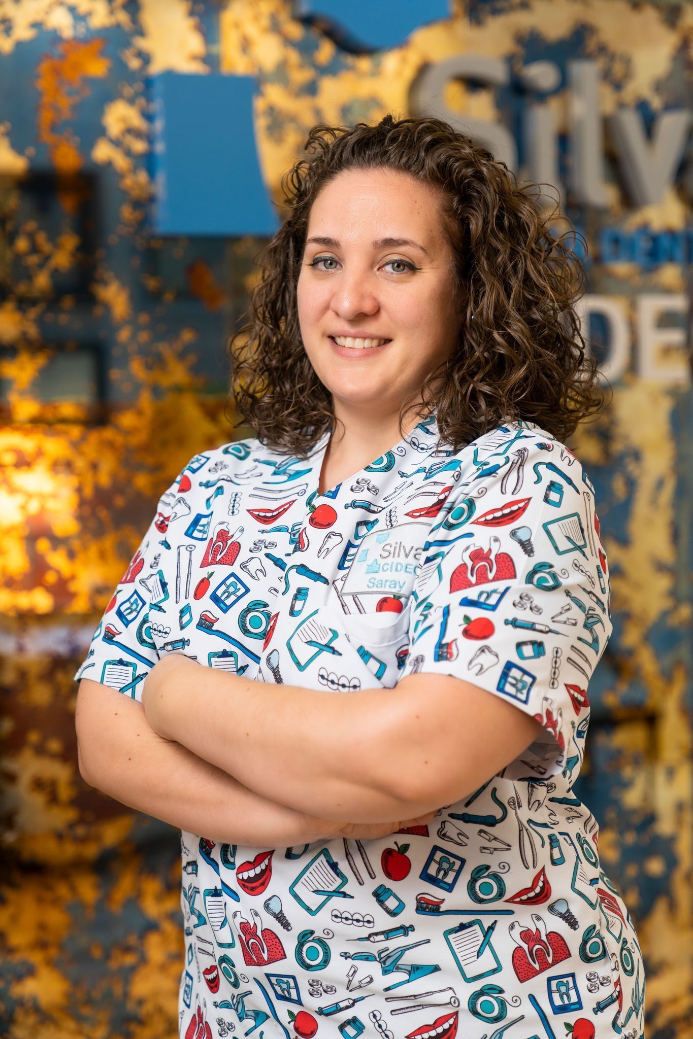 Una mujer con cabello castaño rizado y un cepillo de dientes estampado sonríe y tiene los brazos cruzados.