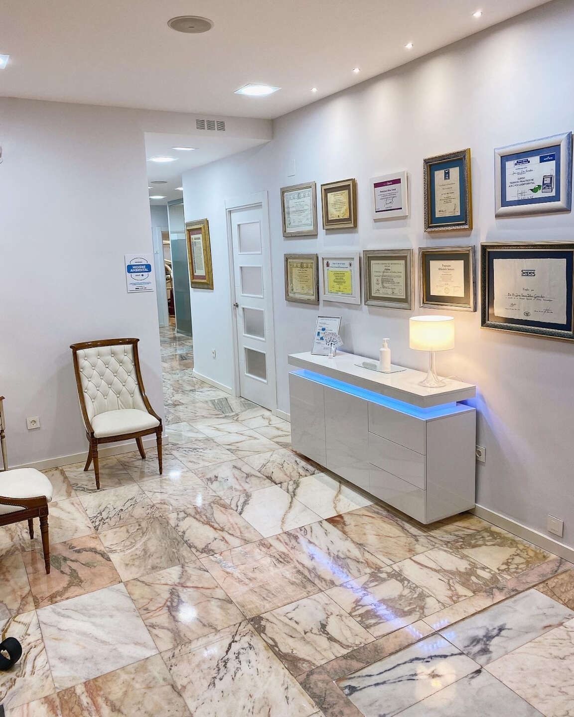 Pasillo en un consultorio dental con pisos de mármol, certificados en la pared y sillas de espera.