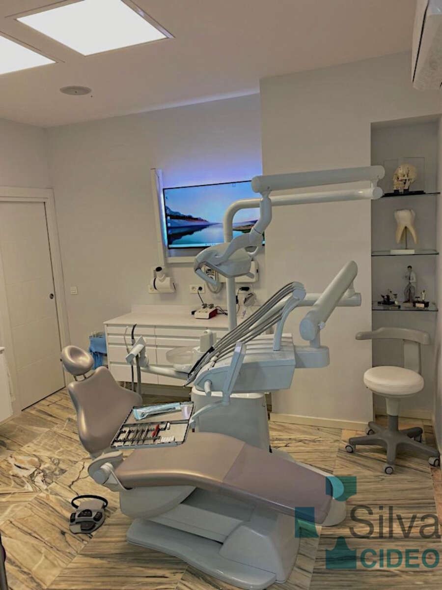 Operario dental con sillón dental, equipo y pantalla de visualización; paredes blancas, tonos neutros y piso.