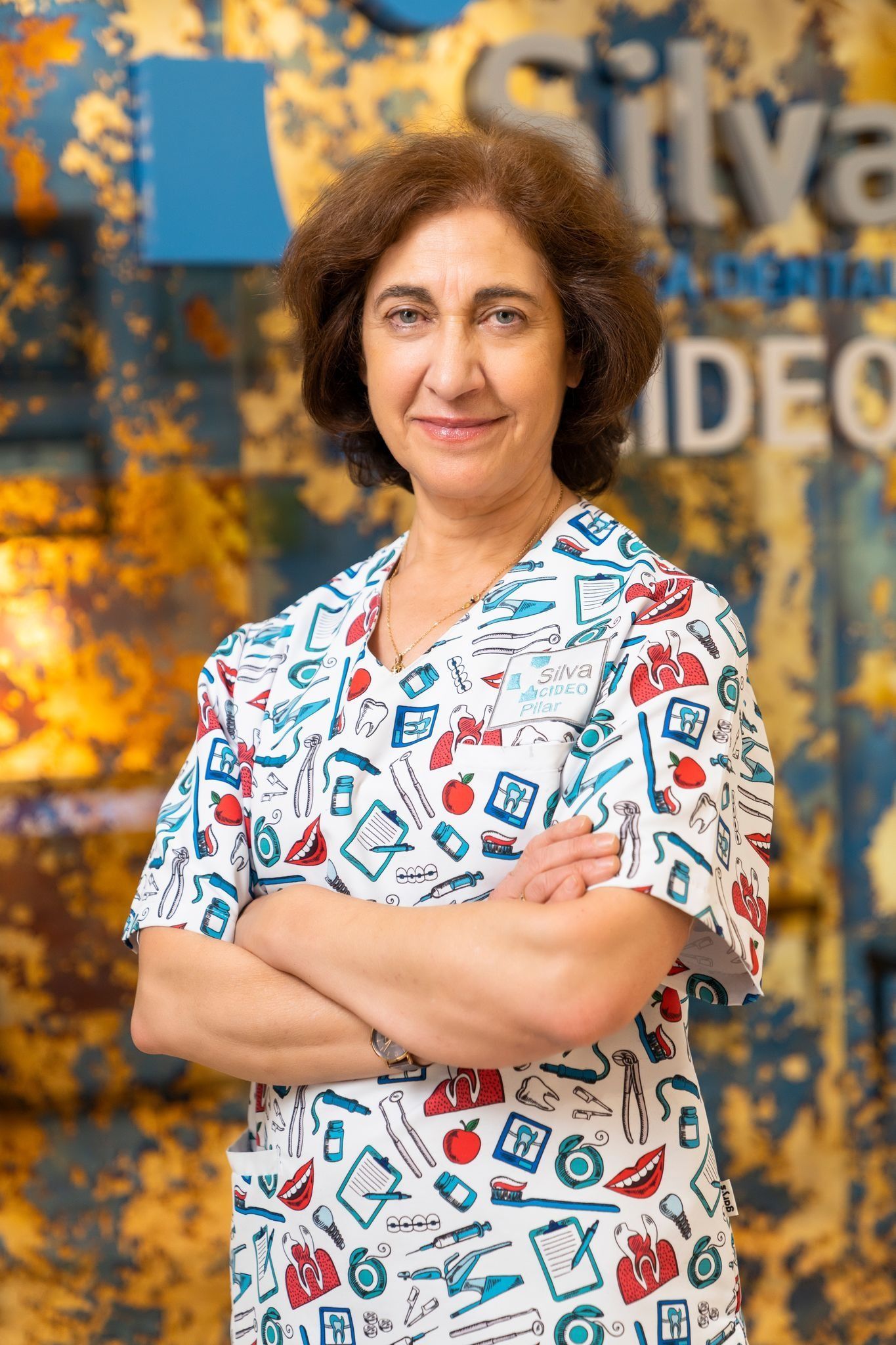 Mujer con cabello castaño rizado con un top estampado, con los brazos cruzados, sonriendo, sobre un fondo borroso y colorido.