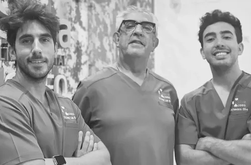 Tres profesionales médicos sonrientes, con uniforme médico. El hombre del centro lleva gafas; los demás tienen los brazos cruzados.