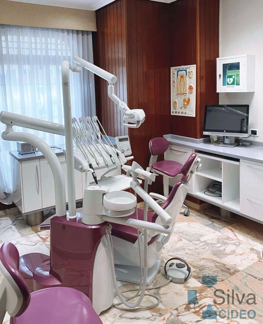 Interior de clínica dental con sillón dental, instrumentos, gabinetes y cuadro anatómico.