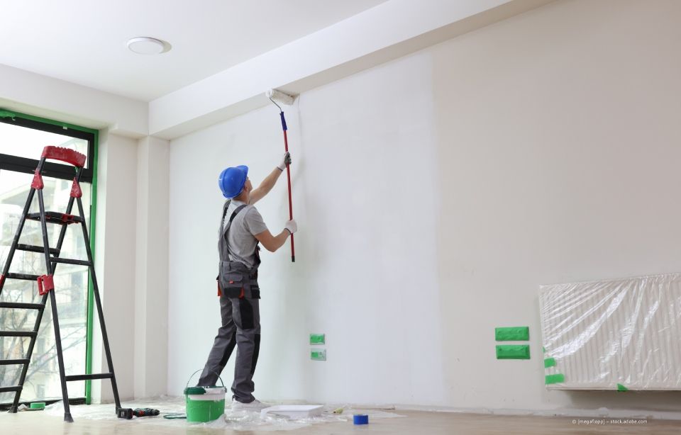Maler mit blauem Hut und Overall trägt weiße Farbe auf eine Wand in einem leeren Raum auf.