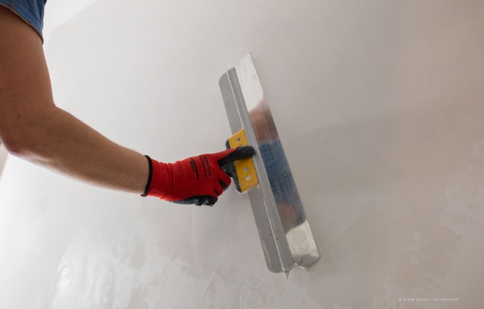 Person mit roten Arbeitshandschuhen glättet weiße Wand mit Metallkelle.